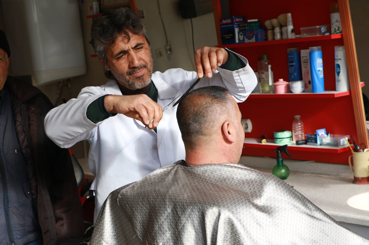 A barber skillfully cuts a customer's hair in a cozy, well-stocked shop.